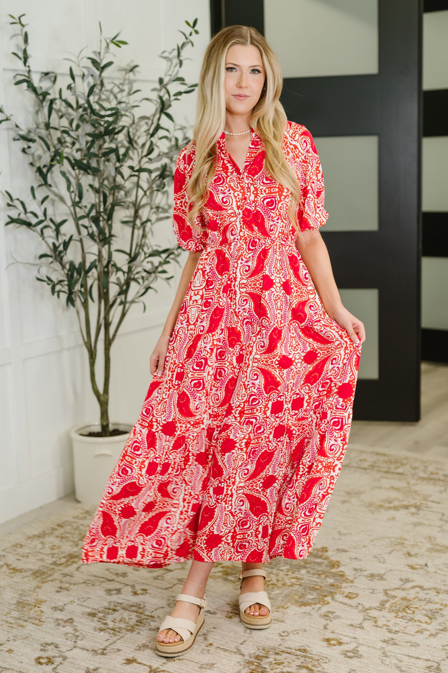 Woman wearing a red patterned dress standing indoors with a plant in the background